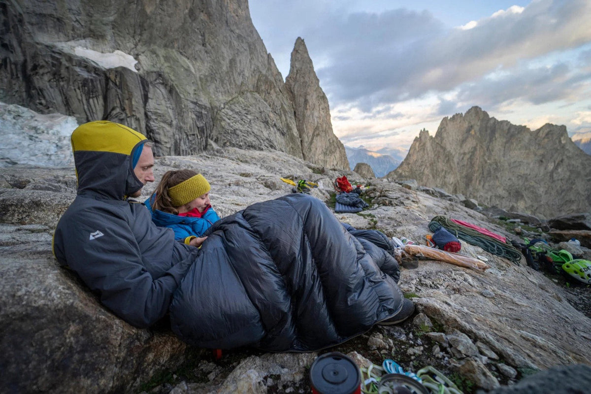 An adult and a child tucked into a Mountain Equipment down sleeping bag at an alpine bivvy, granite peaks behind at dusk