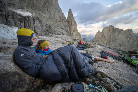 An adult and a child tucked into a Mountain Equipment down sleeping bag at an alpine bivvy, granite peaks behind at dusk