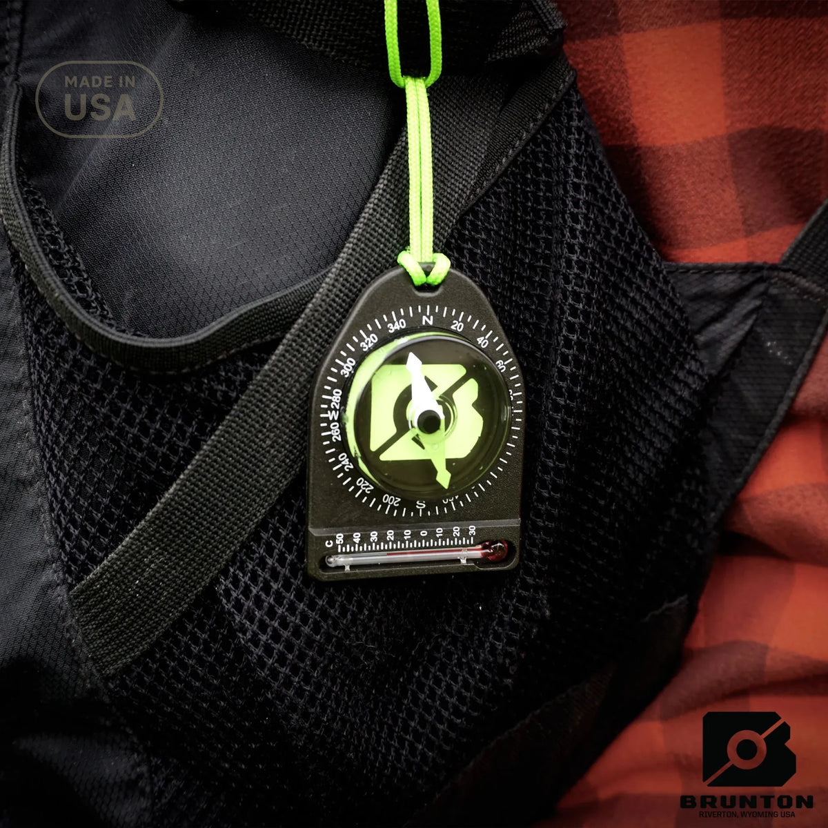 Brunton compass hanging from a backpack strap with a red and black checkered fabric background