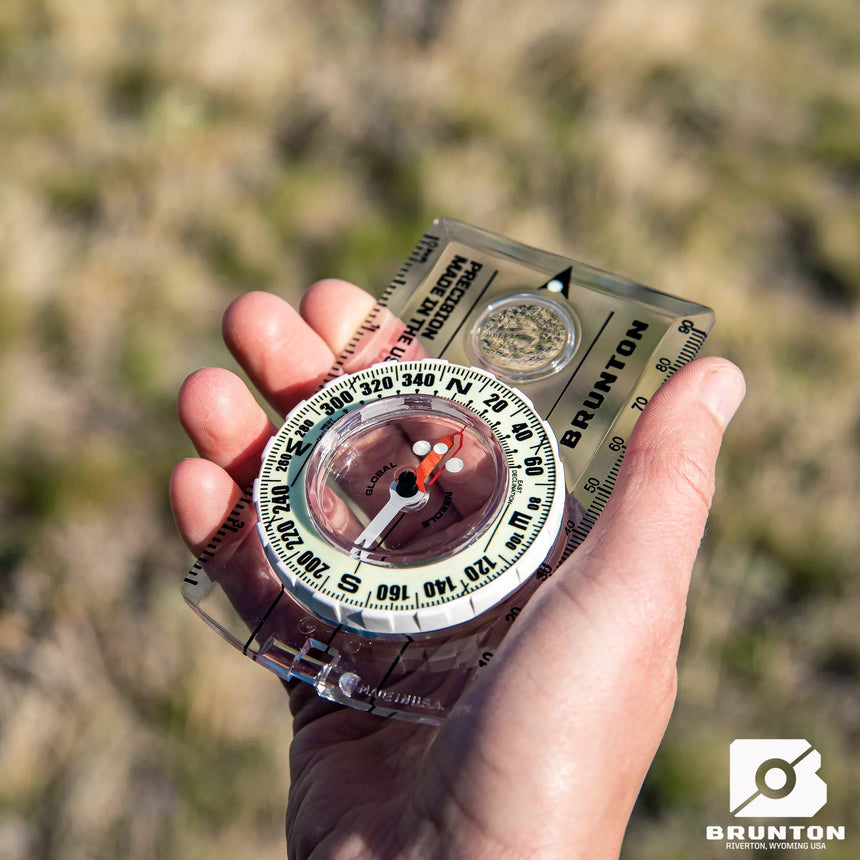 Hand holding a Brunton compass against a blurred natural background