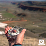 Hand holding a Brunton compass with a scenic landscape in the background