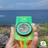 Hand holding a green Brunton compass with ocean and sky in the background