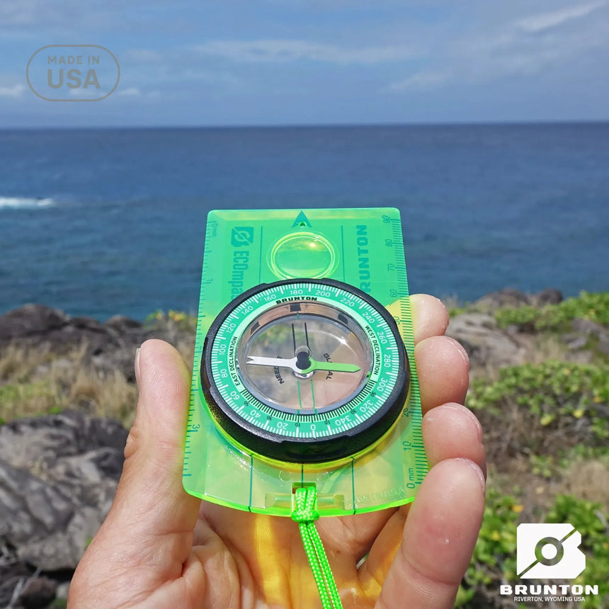 Hand holding a green Brunton compass with ocean and sky in the background